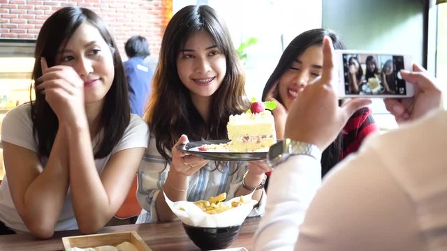 Friends group of Asian people, one male taking photos of three female friends with a strawberry cake at restaurant while girls keep changing poses