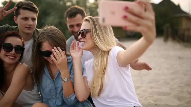 Shot of a group of young friends taking a selfie on the beach. Men and women taking photos sitting on a sand on warm summer evening.