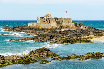 Fototapeta na wymiar The Fort National built by french military architect Vauban on a tidal island, seen from the city of Saint-Malo, France, with the french flag blowing in the wind and rocks in the sea in the foreground