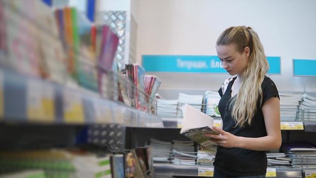 Pretty young woman is regarding sheets of album in a office supplies department. Preparing and shopping for educational year