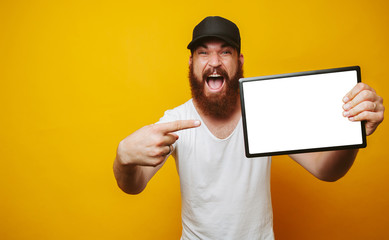 Cheerful amazed happy bearded hipster man in white t-shirt pointing at white screen on tablet at...
