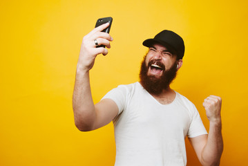 Portrait of a happy young bearded man in white t-shirt looking at mobile phone and celebrating...