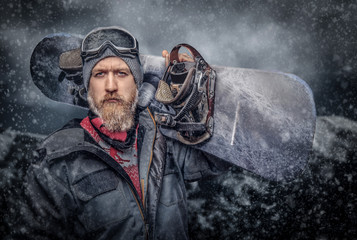 Brutal redhead snowboarder with a full beard in a winter hat and protective glasses dressed in a...