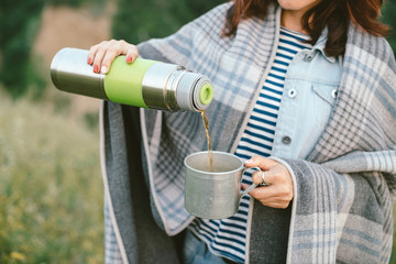 Hands of girl with thermos pours hot tea into mug on meadow on background of trees at misty morning. Girl with red hair in warm blanket in haze.