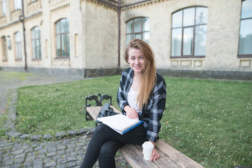 Cute student girl on a bench near the university with books, notebooks and a cup of coffee in his hands looks at the camera and smiles. Student Concept