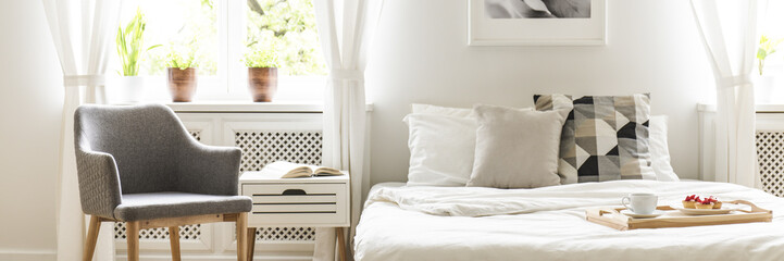 Grey armchair standing by the bedside table with open book in the real photo of white bedroom interior with double bed with cushions and white sheets and fresh plants on windowsill