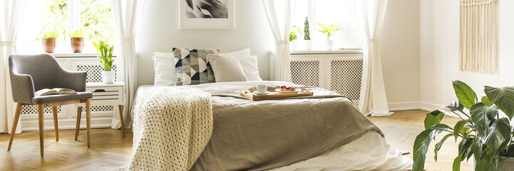 Real photo of bright bedroom interior with king-size bed with wooden tray with breakfast and knit...