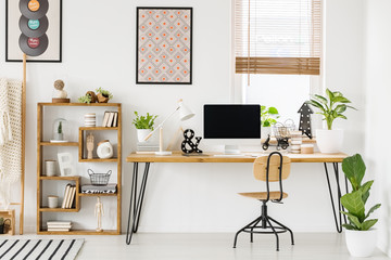 Large industrial desk with a computer by a window, a wooden bookcase and posters on a white wall in...
