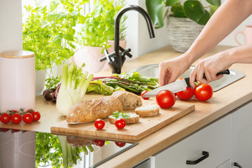 Woman preparing healthy breakfast, cutting a tomato in half and organic herbs and vegetables in a sunny kitchen interior