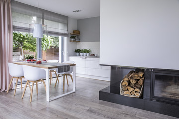 Modern fireplace and wood in a spacious dining room interior with white chairs by a wooden table...