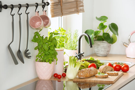 Rye bread on a cutting board, tomatoes and vegetables on a white kitchen interior countertop with Basil herbs in a pink pot