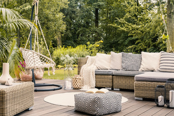 Book on a black and white pouf in the middle of a bright terrace with a rattan corner sofa, hanging...