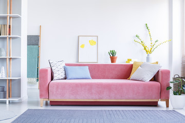 Colorful pillows on a big, velvet, pink sofa in a fun, spacious living room interior with white...