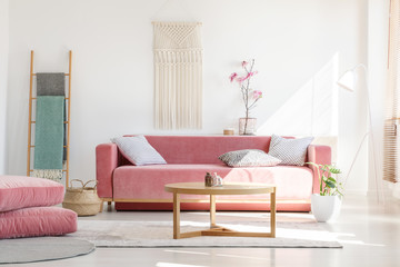 Cozy cushions and stylish textiles in a sunny, feminine living room interior with a pink, velvet sofa and a macrame on a white wall - obrazy, fototapety, plakaty