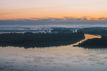 Early blue sky reflected in river water. Riverbank with forest under predawn sky. Cloudy sunrise sky on background. Fog hid trees on island. Colorful morning atmospheric landscape of majestic nature.