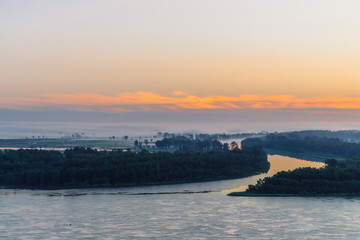 Naklejka premium Early blue sky reflected in river water. Riverbank with forest under predawn sky. Orange stripe in picturesque sky. Fog hid trees on island. Colorful morning atmospheric landscape of majestic nature.