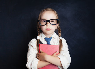 Smart child girl in school uniform clothes with red book on chalk board background. Back to school,...
