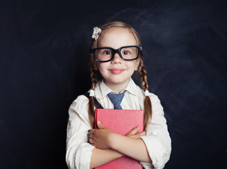Smiling child girl in school uniform clothes with book on chalk board background. Smart kid...