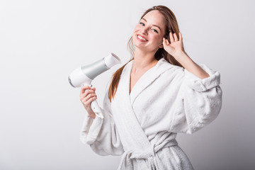  Beautiful red-haired girl in a white bathrobe bathing her hair with a hair dryer on a light background