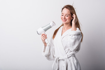  Beautiful red-haired girl in a white bathrobe bathing her hair with a hair dryer on a light background