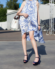 Young leggy woman blogger posing outdoors in a fashionable white dress with a blue pattern with a large white hand bag in the city park. Part of the body.