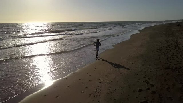 Aerial footage - Jogger on a lonely sand beach runs at sunset with light reflection on the sea surface