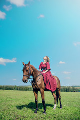 Beautiful girl in a long red dress riding a brown horse against a blue sky. The girl looked up and looked at the sky.