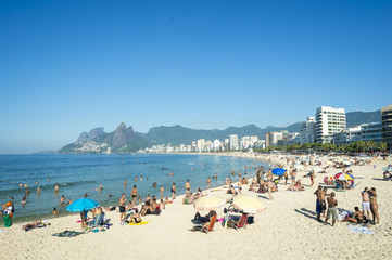 Fototapeta na wymiar Rio residents, known as cariocas, relax at the Arpoador section of Ipanema Beach with the city skyline and Two Brothers Mountain in the background.