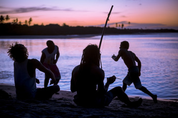 Silhouettes of four unrecognizable capoeiristas sat on the beach paying berimbau and drum...