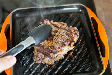 Cooking meat on cast iron grill pan indoors at home. Person turning ribeye steak on other side with tongs