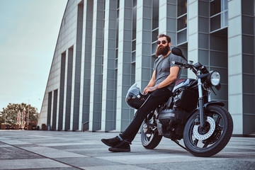 Brutal bearded male in a gray t-shirt and black pants holds a helmet sitting on his custom-made...