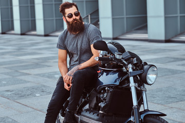 Brutal bearded male in sunglasses dressed in a gray t-shirt and black pants sitting on his custom-made retro motorcycle against a skyscraper.