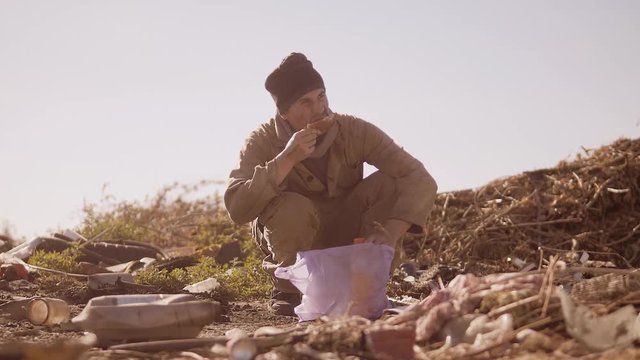 portrait of a dirty homeless hungry man in a dump eating for food in the package with walking goes looking for food slow motion video. homeless dirty man roofless person looking for food in a dump