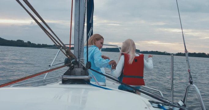 Young adult mid-30s Caucasian female and her daughter sailing on yacht, sunset in the background. 4K UHD 60 FPS SLO MO