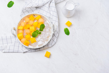 Thai dessert food style : Top view of homemade sago with coconut milk and topping with mango cut into squares and cantaloupe ball in a white bowl  and placed on a marble table with a tablecloth plaid.