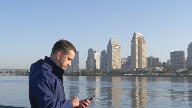 Man sms texting using app on smart phone at bacground downtown San Diego. Young man sends a sms text message on phone. Young man texting on smartphone standing on City. Traveler writes a sms message.