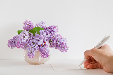 Fresh bouquet of purple lilac blossoms in the vase. Woman's hand with pen writing on the white blank greeting card on the table. Empty place for inspirational, emotional, sentimental text or quote.