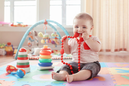 Portrait Of Cute Adorable Blond Caucasian Smiling Child Boy With Blue Eyes Sitting On Floor In Kids Children Room. Little Baby Playing With Teething Toys Jewelry. Early Education Development Concept.