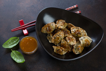 Fried wontons with sesame seeds served in a black bowl with dipping sauce, horizontal shot on a brown metal background
