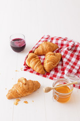French style breakfast, a white wooden table with glass of fresh beetroot smoothie, croissants and glass bowl of honey, few croissants placed on the red and white french checkered fabric.