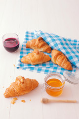 French style breakfast, a white wooden table with glass of fresh beetroot smoothie, croissants and glass bowl of honey, few croissants placed on the blue, turquoise and white checkered fabric.