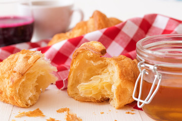 French style breakfast, a white wooden table with glass of fresh beetroot smoothie, croissants and glass bowl of honey, few croissants placed on the red and white checkered fabric. Close up.