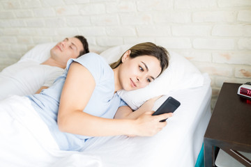 Woman Secretly Using Smartphone While Lying By Boyfriend On Bed