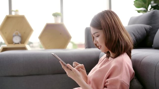 Asian woman using tablet while lying on home sofa in her living room. Happy female use tablet for video call with her friends or boyfriend at home.