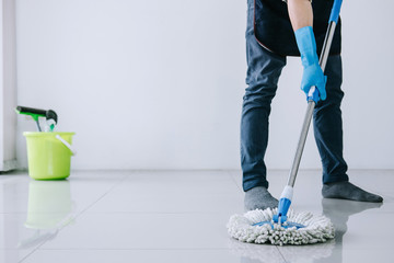 Fototapeta premium Husband housekeeping and cleaning concept, Happy young man in blue rubber gloves wiping dust using mop while cleaning on floor at home