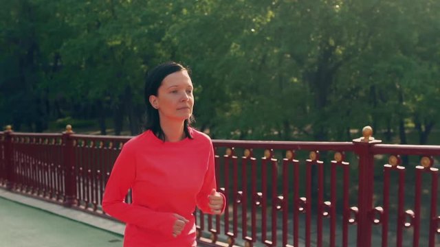 adult woman jogging outdoors slow motion. female wearing red sportswear long sleeve t-shirt running in park with green trees