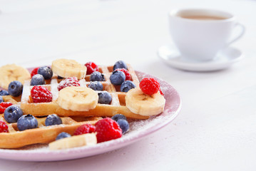 Cup of coffee and belgian waffles with blueberries, raspberries, bananas and sugar powder on white background. Concept of tasty and healthy food.