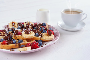 Cup of coffee and tasty belgian waffles with blueberries, raspberries, bananas and sugar powder are filled with liquid chocolate.