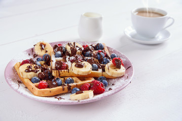 Cup of coffee and tasty belgian waffles with blueberries, raspberries, bananas and sugar powder are filled with liquid chocolate.