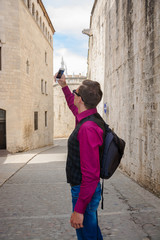A young tourist with a backpack takes pictures on a mobile phone in the center of Girona near the Basilica of Saint Felix
